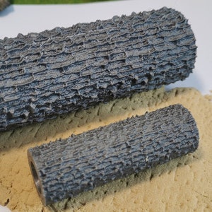 May include: Two grey textured cylindrical moulds for creating bark-like patterns on crafts. The moulds are on a bed of tan sand.