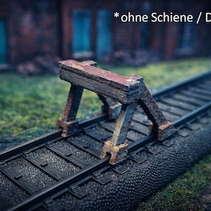 Op de afbeelding: Een miniatuur spoorweg met een verweerde houten vakwerkbrug. De brug is bruin en de rails zijn zwart. De achtergrond is wazig, met een bakstenen gebouw en groen gras zichtbaar. De tekst "* ohne Schiene / Deko" staat in het midden bovenaan.