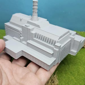 Può includere: Modello grigio stampato in 3D di un edificio con una struttura cilindrica alta sulla parte superiore. Il modello è tenuto in mano su uno sfondo verde e blu.