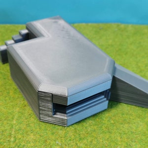 Könnte beinhalten: Ein graues Betonbunkermodell mit einem kleinen Eingang und einer Rampe, die zu ihm führt. Das Modell steht auf einer grünen Grasfläche.