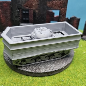 Könnte beinhalten: Ein graues Miniatur-Panzermodell in einem Sarg-förmigen Behälter. Der Panzer ist mit einem Turm und Ketten ausgestattet, und der Behälter hat ein geschichtetes Design. Das Modell steht auf einer kreisförmigen Basis, mit einem Hintergrund eines zerstörten Gebäudes.