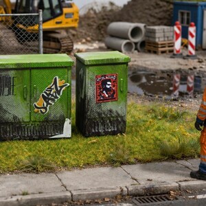 Pode incluir: Duas caixas verdes de utilidade pública com arte de grafite estão em uma área gramada perto de um canteiro de obras. Dois trabalhadores com roupas de segurança laranja e azul estão na calçada, observando a cena. Uma escavadeira amarela e outros materiais de construção estão ao fundo.