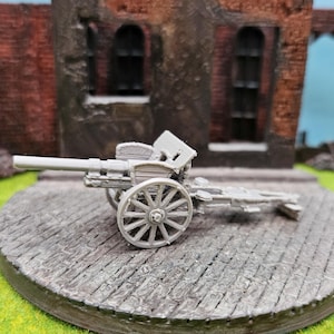 Könnte beinhalten: Ein graues Miniaturmodell einer Artilleriekanone mit Speichenrädern, das auf einer kreisförmigen Plattform mit einem ziegelartigen Muster steht. Der Hintergrund zeigt ein Diorama eines zerstörten Backsteingebäudes und grünes Gelände.