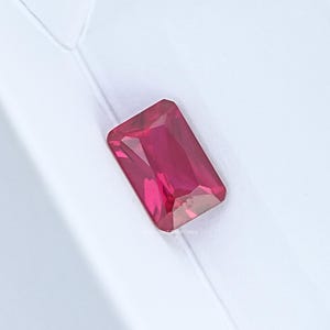 Puede incluir: Una gema rectangular de color rojo rubí con una superficie pulida. La piedra preciosa se encuentra sobre un fondo blanco, resaltando su color vibrante y claridad. El corte de la piedra es de esmeralda clásico.