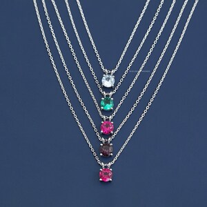 May include: Five silver necklaces with different colored gemstones. The necklaces are layered and feature a light blue, green, pink, burgundy, and pink gemstone.