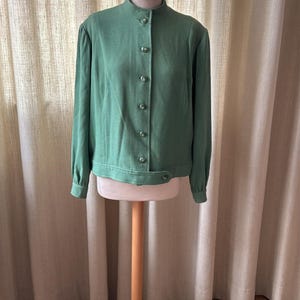 Può includere: Giacca verde in stile vintage con collo alto e chiusura a bottoni. La giacca a maniche lunghe è esposta su un manichino di legno. La giacca ha una vestibilità sartoriale e un design classico, adatta a varie occasioni.