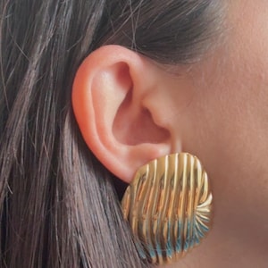 Può includere: Orecchini quadrati color oro con superficie strutturata e scanalata. Gli orecchini sono attaccati a un orecchio, con capelli castano scuro visibili sullo sfondo. Gli orecchini hanno un'estetica vintage.