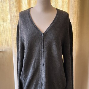 May include: A gray knitted cardigan with a v-neck and button closure. The cardigan is on a wooden mannequin.