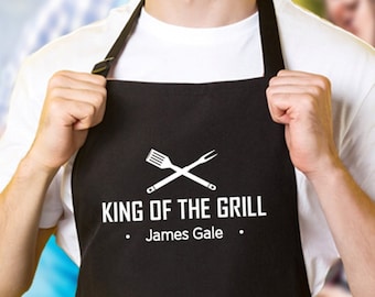 Könnte beinhalten: Eine schwarze Schürze mit einer weißen Grafik aus gekreuztem Pfannenwender und Gabel über dem Text KING OF THE GRILL und dem Namen James Gale. Die Schürze verfügt über eine große Vordertasche und verstellbare Nackenbänder für das Grillen.