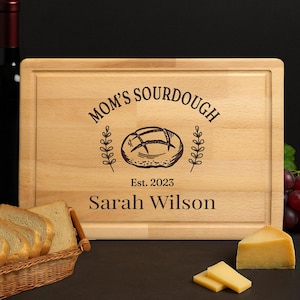 May include: A rectangular wooden cutting board with the text "MOM'S SOURDOUGH" and "Est. 2023" above the name "Sarah Wilson". The board features a bread illustration and is surrounded by a decorative border. Slices of bread and cheese are in the foreground.