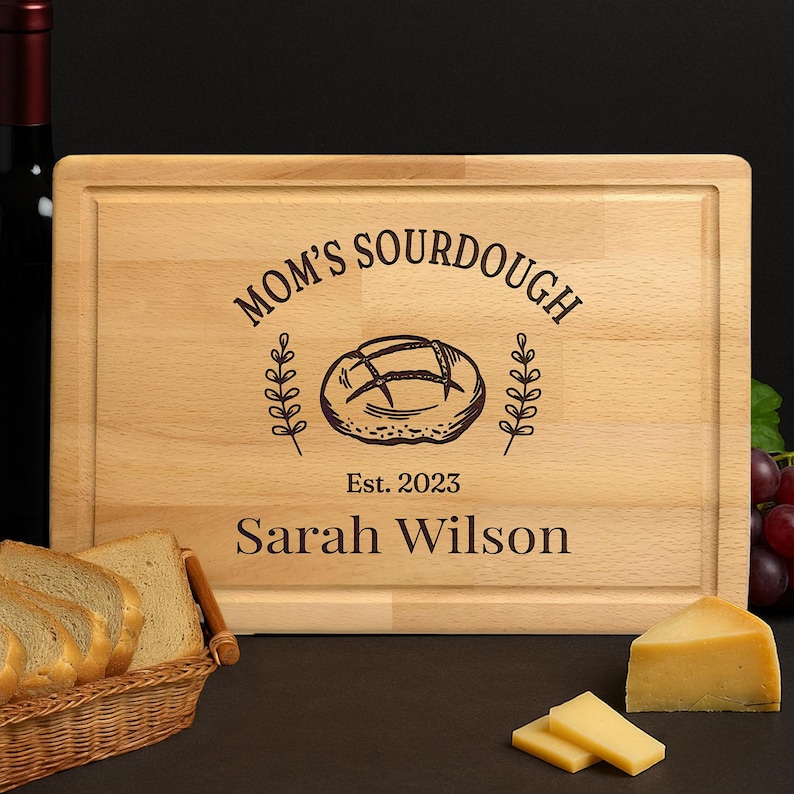 May include: A rectangular wooden cutting board with the text "MOM'S SOURDOUGH" and "Est. 2023" above the name "Sarah Wilson". The board features a bread illustration and is surrounded by a decorative border. Slices of bread and cheese are in the foreground.