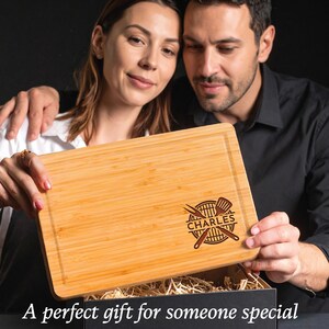 Puede incluir: Tabla de cortar rectangular de bamb&uacute; con un dise&ntilde;o grabado en marr&oacute;n oscuro que presenta una parrilla, pinzas y el nombre "CHARLES". La tabla est&aacute; siendo sostenida por una pareja. El texto en la parte inferior dice "Un regalo perfecto para alguien especial."