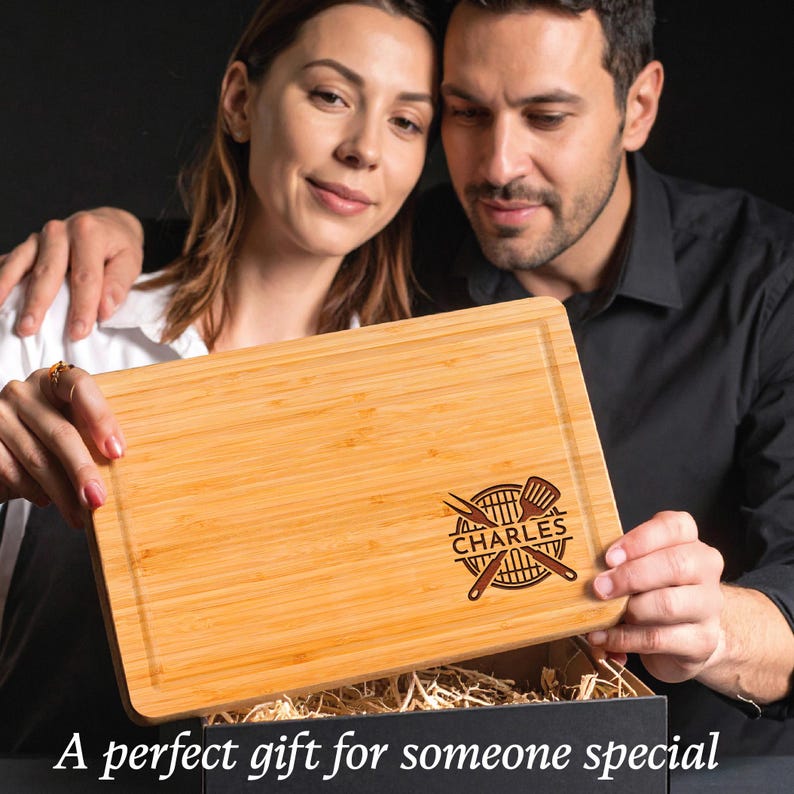 Puede incluir: Tabla de cortar rectangular de bamb&uacute; con un dise&ntilde;o grabado en marr&oacute;n oscuro que presenta una parrilla, pinzas y el nombre "CHARLES". La tabla est&aacute; siendo sostenida por una pareja. El texto en la parte inferior dice "Un regalo perfecto para alguien especial."