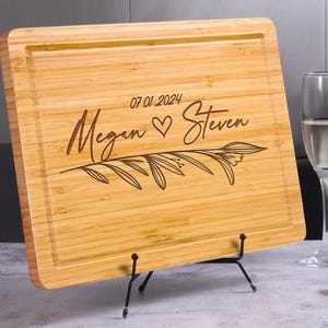 May include: A natural bamboo cutting board with a personalized engraved message. The message reads "Megan & Steven" with a heart symbol and the date "07 01 2024". The board is displayed on a black metal stand.