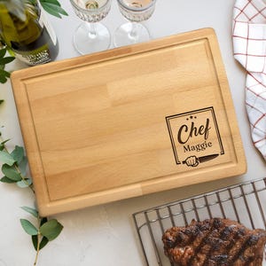 Puede incluir: Una tabla de cortar rectangular de madera con ranura para jugos. La tabla presenta el texto "Chef Maggie" en un dise&ntilde;o enmarcado con una ilustraci&oacute;n de cuchillo. La tabla est&aacute; rodeada de una botella de vino, copas y un bistec.