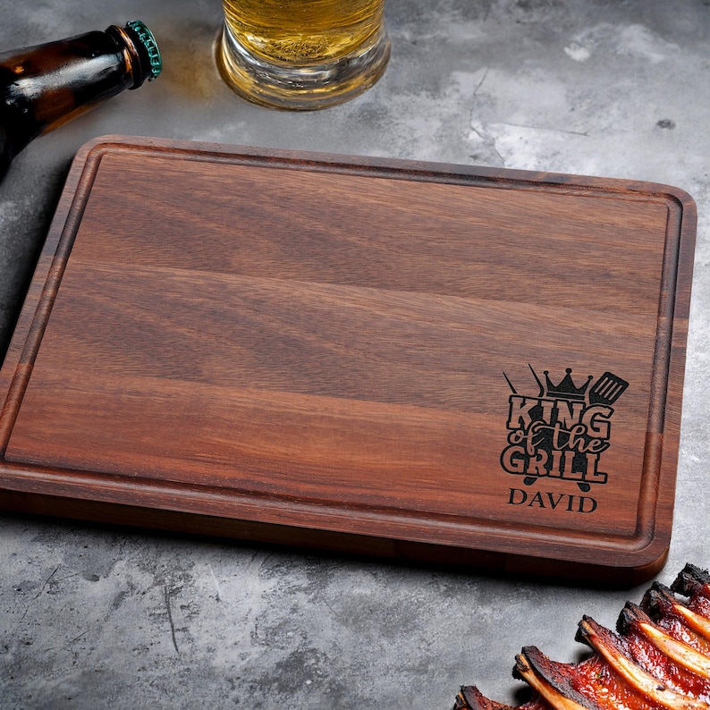 Puede incluir: Tabla de cortar rectangular de madera marr&oacute;n oscuro con borde rebajado. Presenta el texto "KING of the GRILL" con una corona y utensilios de parrilla, y el nombre "DAVID" debajo. Una botella y un vaso de cerveza se ven en el fondo. Aproximadamente 30 cm x 20 cm.