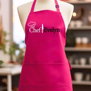 May include: A bright pink apron featuring a white chef hat icon, a white knife graphic, and the black text "Chef Evelyn" printed on the chest. The apron includes a neck strap with an adjustable black buckle and a large front pocket across the waist.
