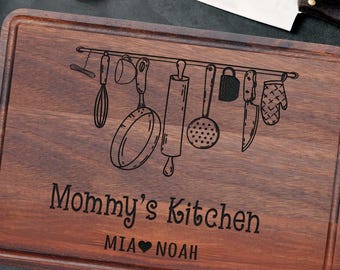 Könnte beinhalten: Ein rechteckiges, dunkelbraunes Holzschneidebrett mit einer Lasergravur verschiedener Küchenutensilien, die an einer Leiste hängen. Der Text Mommy's Kitchen und MIA Herz NOAH ist unter der Utensiliengrafik in Schwarz eingraviert.