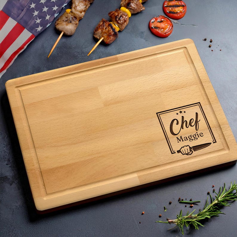 Puede incluir: Tabla de cortar rectangular de madera clara con ranura para jugos. La tabla tiene el texto "Chef Maggie" en un marco cuadrado con un gr&aacute;fico de cuchillo. Borde marr&oacute;n oscuro. Brochetas de carne a la parrilla y tomates en el fondo.