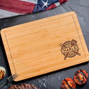 Puede incluir: Una tabla de cortar rectangular de bamb&uacute; con un borde oscuro y una ranura tallada. La tabla presenta un dise&ntilde;o tem&aacute;tico de barbacoa con el nombre "CHARLES" y utensilios para asar. El fondo incluye tomates a la parrilla y una bandera estadounidense.