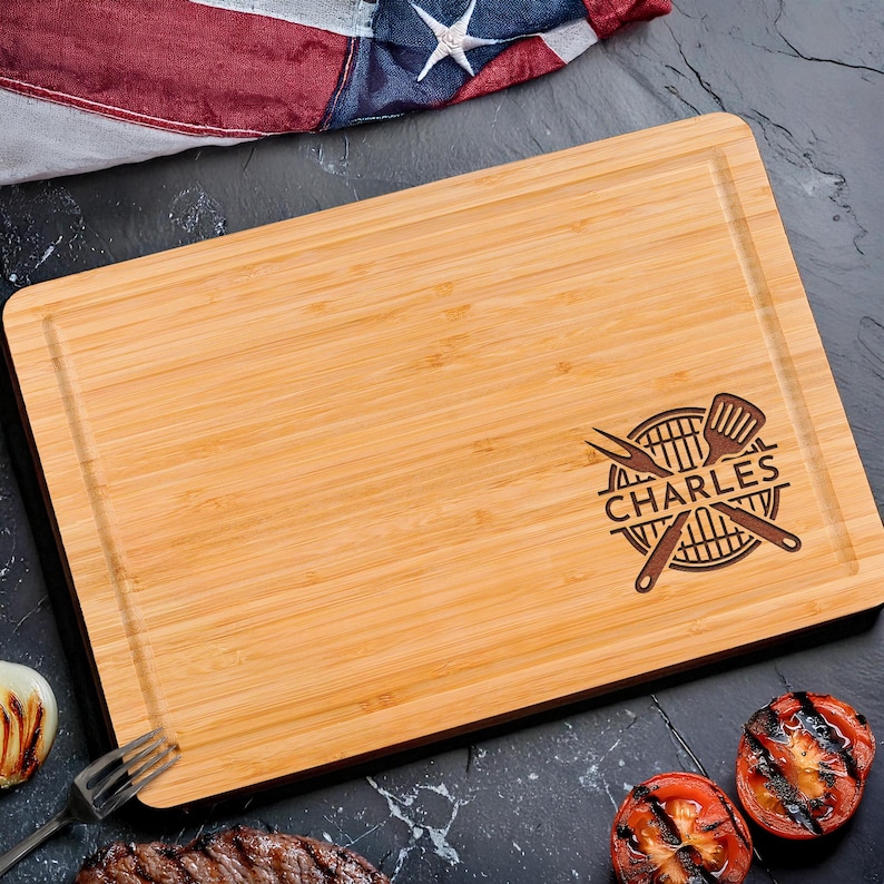 Puede incluir: Una tabla de cortar rectangular de bamb&uacute; con un borde oscuro y una ranura tallada. La tabla presenta un dise&ntilde;o tem&aacute;tico de barbacoa con el nombre "CHARLES" y utensilios para asar. El fondo incluye tomates a la parrilla y una bandera estadounidense.