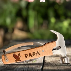 Może przedstawiać: Młotek wielofunkcyjny z drewnianą rączką. Na rączce wygrawerowano napis "PAPA" i grafikę orła. Narzędzie zawiera głowicę młotka, n&oacute;ż i inne narzędzia. Narzędzie wykonane jest z drewna i metalu.