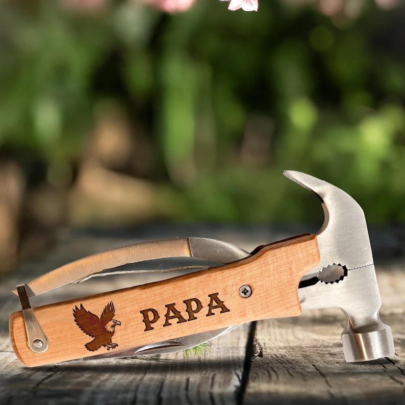 Może przedstawiać: Młotek wielofunkcyjny z drewnianą rączką. Na rączce wygrawerowano napis "PAPA" i grafikę orła. Narzędzie zawiera głowicę młotka, n&oacute;ż i inne narzędzia. Narzędzie wykonane jest z drewna i metalu.