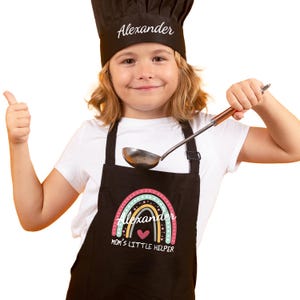 May include: A black apron with a rainbow design and the name "Alexander" in white script. The apron also features the text "MOM'S LITTLE HELPER" and a heart. A black chef's hat with "Alexander" in white script is also visible.