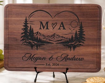 Könnte beinhalten: Ein rechteckiges Schneidebrett aus dunkelbraunem Holz mit einer Lasergravur von Bergen, Tannen und einem See. Die Gravur enthält die Buchstaben M und A, getrennt durch ein Herz, die Namen Megan & Andrew sowie den Text Est. 2026.