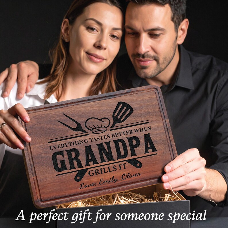 May include: A rectangular wooden cutting board with a dark brown finish. The board is engraved with the words "EVERYTHING TASTES BETTER WHEN GRANDPA GRILLS IT" and includes a chef's hat, spatula, and fork design. The names "Emily, Oliver" are also engraved.