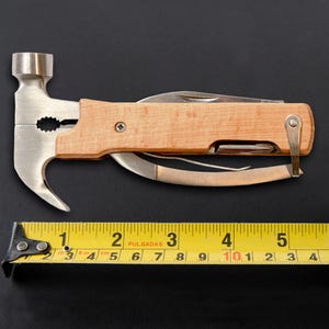 K&ouml;nnte beinhalten: Ein Multitool mit Hammerkopf, Zange, Messer und anderen Werkzeugen. Der Griff ist aus hellem Holz gefertigt. Ein gelbes Ma&szlig;band mit schwarzen Markierungen liegt neben dem Werkzeug. Das Ma&szlig;band zeigt Messungen in Zentimetern an.