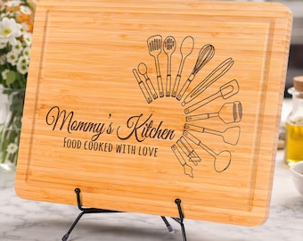 Könnte beinhalten: Ein rechteckiges Schneidebrett aus hellem Bambus mit einer Lasergravur verschiedener Küchenutensilien in Fächerform. Der Text lautet Mommy's Kitchen und FOOD COOKED WITH LOVE. Das Brett steht auf einem schwarzen Metallständer.