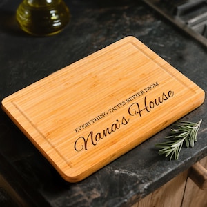 May include: A rectangular bamboo cutting board with a juice groove. The text "EVERYTHING TASTES BETTER FROM Nana's House" is engraved on the board. The board is light brown and sits on a dark countertop.