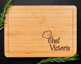 Könnte beinhalten: Ein rechteckiges Schneidebrett aus Bambus mit einer Saftrille am Rand. Das Brett ist mit einer Lasergravur eines Kochmützen-Symbols über dem Text "Chef Victoria" in dunkelbrauner Schrift versehen, akzentuiert durch grüne Rosmarinzweige.
