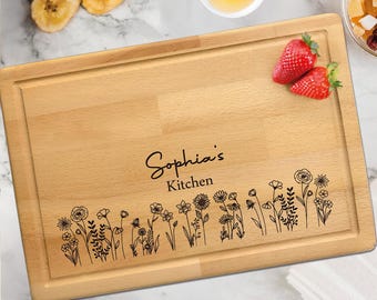 Könnte beinhalten: Ein rechteckiges Schneidebrett aus hellem Holz mit einer Saftrille am Rand. Das Brett ist mit dem Text Sophia's Kitchen über einer detaillierten schwarzen Strichzeichnung verschiedener Wildblumen und Stängel graviert.