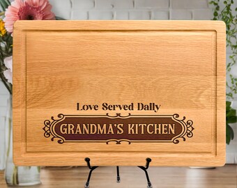 Könnte beinhalten: Ein rechteckiges, hellbraunes Holzschneidebrett mit einer Saftrille am Rand. Das Brett zeigt den eingravierten Text "Love Served Daily" über einem dekorativen Rahmen, der den Text "GRANDMA'S KITCHEN" in dunkler Schrift enthält.
