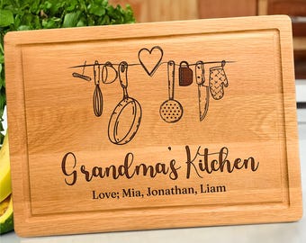 Könnte beinhalten: Ein rechteckiges Schneidebrett aus hellem Holz mit einer Saftrille. Das Brett zeigt eine lasergravierte Illustration von Küchenutensilien, die an einer Linie mit einem Herz hängen, sowie den Text "Grandma's Kitchen" und "Love; Mia, Jonathan, Liam".