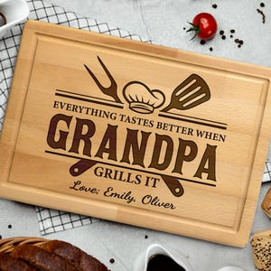May include: A rectangular wooden cutting board with a carved border. The text "EVERYTHING TASTES BETTER WHEN GRANDPA GRILLS IT" is etched into the wood, along with a chef's hat, fork, and spatula. The names "Love; Emily, Oliver" are also etched.