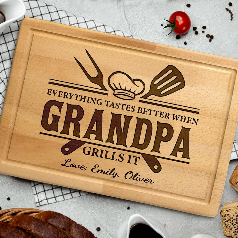 May include: A rectangular wooden cutting board with a carved border. The text "EVERYTHING TASTES BETTER WHEN GRANDPA GRILLS IT" is etched into the wood, along with a chef's hat, fork, and spatula. The names "Love; Emily, Oliver" are also etched.