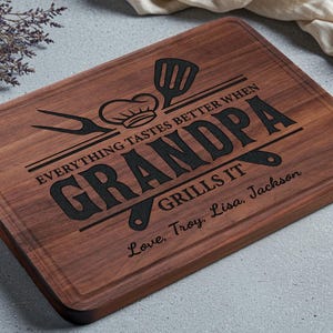 May include: A rectangular wooden cutting board with a dark brown finish. The board features engraved text that reads "EVERYTHING TASTES BETTER WHEN GRANDPA GRILLS IT" along with a chef's hat, spatula, and other grilling tools. The text "Love, Troy, Lisa, Jackson" is also engraved.