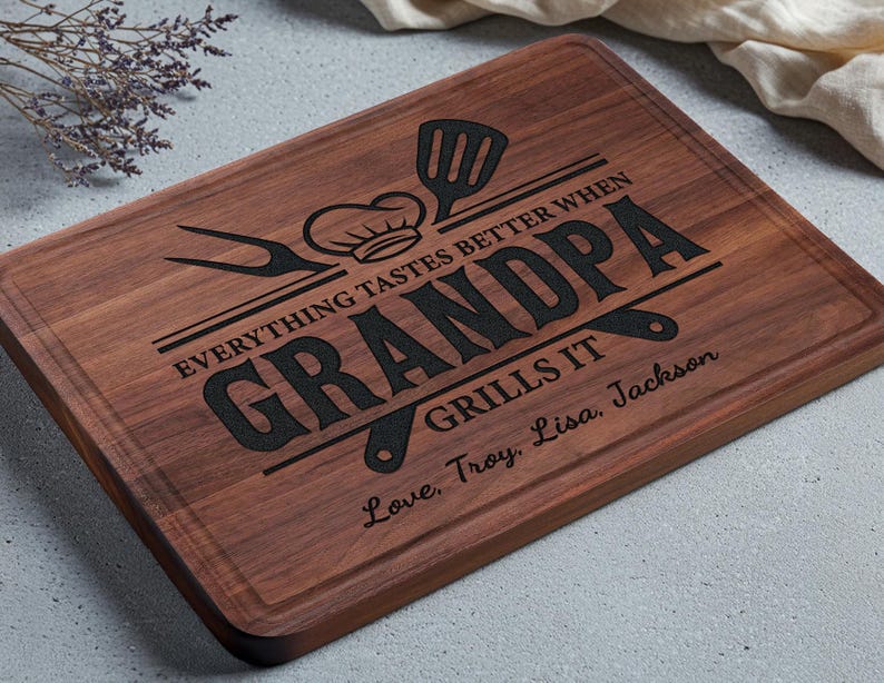 May include: A rectangular wooden cutting board with a dark brown finish. The board features engraved text that reads "EVERYTHING TASTES BETTER WHEN GRANDPA GRILLS IT" along with a chef's hat, spatula, and other grilling tools. The text "Love, Troy, Lisa, Jackson" is also engraved.
