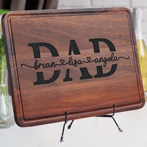 May include: A rectangular wooden cutting board with a dark brown finish. The word "DAD" is prominently displayed in large, black letters, with the names "brian", "lisa", and "angela" below. The board is supported by a black metal stand.