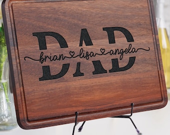 May include: A rectangular wooden cutting board with a dark brown finish. The word "DAD" is prominently displayed in large, black letters, with the names "brian", "lisa", and "angela" below. The board is supported by a black metal stand.