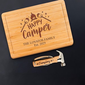 May include: A rectangular bamboo cutting board with a carved design that reads "Happy Camper" with a campfire and marshmallow graphic. Below the text is "THE LOVATOS FAMILY Est. 2025". A camping multi-tool with a hammer is below the cutting board.