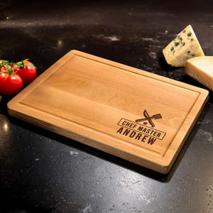 Puede incluir: Tabla de cortar rectangular de madera con borde empotrado. La tabla est&aacute; grabada con el texto "CHEF MASTER ANDREW" y un s&iacute;mbolo de cuchillo y tenedor cruzados. Tomates y queso en el fondo.