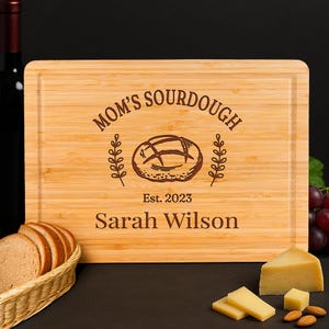 May include: A rectangular bamboo cutting board with the words "MOM'S SOURDOUGH" and "Est. 2023 Sarah Wilson" engraved on it. The board features a bread loaf illustration and is surrounded by bread, cheese, and almonds.
