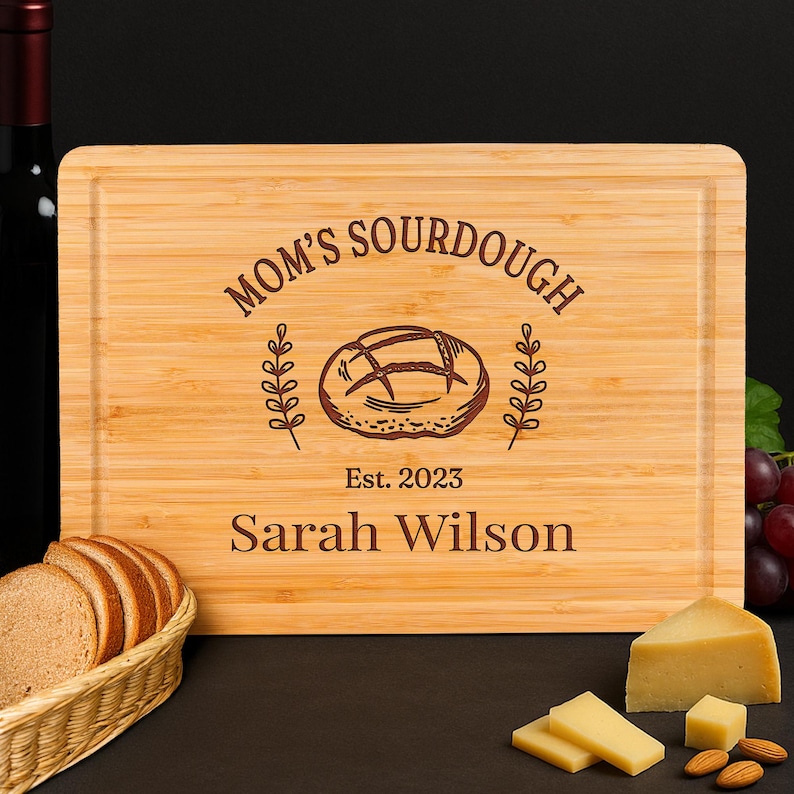 May include: A rectangular bamboo cutting board with the words "MOM'S SOURDOUGH" and "Est. 2023 Sarah Wilson" engraved on it. The board features a bread loaf illustration and is surrounded by bread, cheese, and almonds.