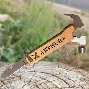 May include: A multi-tool with a wooden handle featuring a hammer, knife, and other tools. The handle is engraved with the name "ARTHUR" and a wrench and screwdriver icon. The tool is resting on a wooden surface.