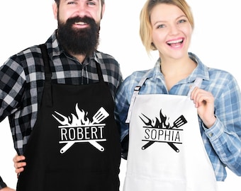 Könnte beinhalten: Eine schwarze Schürze mit dem Namen ROBERT und einer weißen Grafik aus Grillgabel, Pfannenwender und Flammen sowie eine weiße Schürze mit dem Namen SOPHIA und einer schwarzen Grafik aus Grillgabel, Pfannenwender und Flammen. Beide haben Taschen.