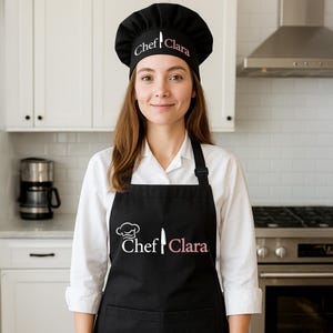 May include: A black chef's hat and apron set with the name "Chef Clara" in pink script. The hat has a knife graphic. The apron has a chef hat and knife graphic. The person is wearing a white shirt.